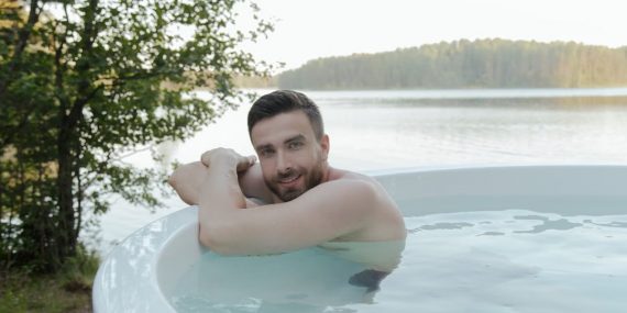 Shirtless man enjoying relaxation in hot tub by scenic lake, smiling and embracing leisure outdoors.